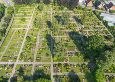 Luftaufnahme eines weitläufigen, gepflegten Friedhofsgeländes mit angelegten Grabfeldern, Gehwegen und schattenspendendem Baumbestand