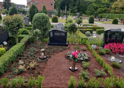 Sorgfältig gepflegte Grabstätte auf einem Friedhof mit dunklem Grabstein, rötlicher Mulchabdeckung, niedrigen Hecken, Grabkerze und rosa Blumenstrauß