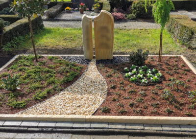 Moderne, harmonisch angelegte Grabstätte mit zweigeteiltem Sandstein-Grabstein, geschwungenem Hellkiespfad, zwei kleinen Bäumchen und abwechslungsreicher Bepflanzung