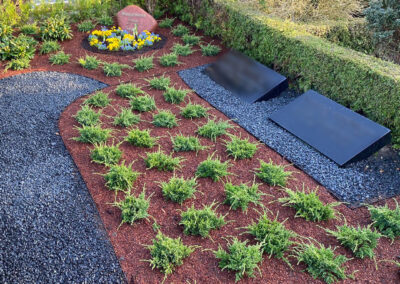 Liebevoll gestaltete Grabstätte mit symmetrischer Bodendecker-Bepflanzung, dunklem Kiesweg, rötlicher Rindenmulch-Abdeckung und gelb-blauen Blumenakzenten um einen Gedenkstein